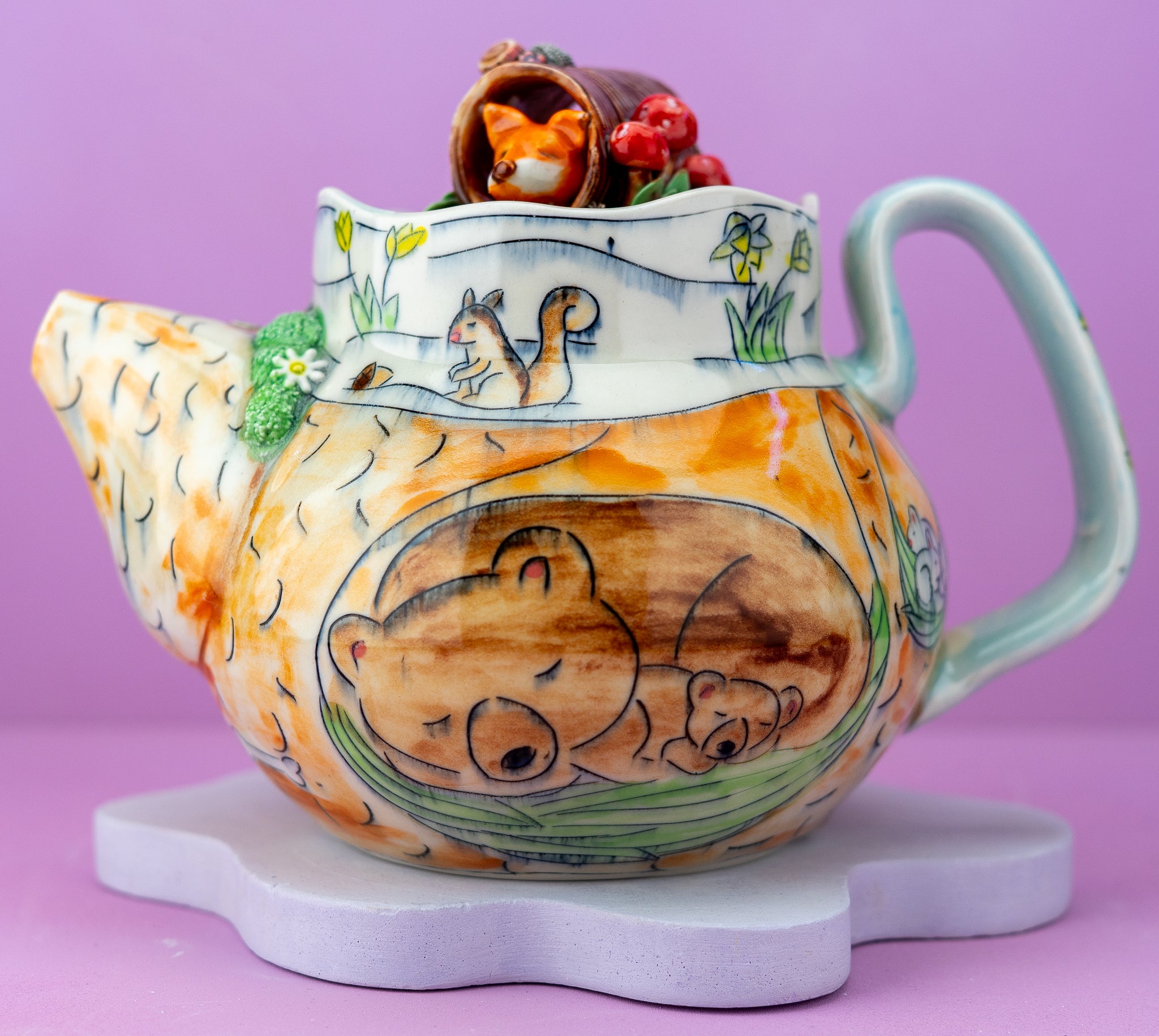 A ceramic teapot with whimsical woodland animal illustrations, including a sleeping bear on the front, a squirrel on the rim, and a fox in a hat on top. The teapot has a light blue handle and spout, a warm orange and brown body, and is displayed on a white cloud-shaped stand against a solid lavender background.